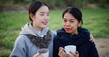 Handheld selective focus shot, Two Asian women standing talking in a garden drinking coffee and enjoying the view in front of them on a cold morning. Winter activities. video