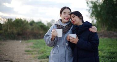Handheld shot, Two Asian women standing talking in a garden holding cups of coffee and enjoying the view in front of them on a cold morning. video