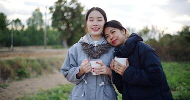 Handheld shot, Two Asian women talking in a garden holding cups of coffee and enjoying the view in front of them on a cold morning. Winter activities. video