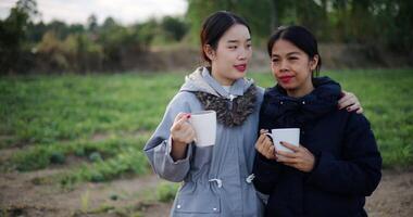Handheld selective focus shot, Two Asian women standing talking in a garden holding cups of coffee and enjoying the view in front of them on a cold morning. Winter activities. video