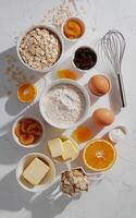 A variety of baking ingredients, including oats, flour, eggs, butter, and dried fruits, are arranged neatly on a white surface. photo