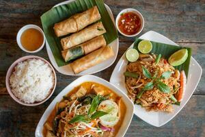An assortment of vibrant Thai dishes, including spring rolls, rice, and a flavorful noodle dish, is beautifully arranged on a rustic wooden table. photo