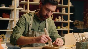 Working artist sculptor Caucasian man guy potter craftsperson ceramist small business owner male in apron making clay sculpt adding texture use tool shaping pottery ceramic artwork at workshop studio video