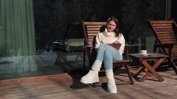 Cozy winter morning finds a woman writer relaxing on a wooden porch, sipping a hot beverage and making notes in her notebook, bathed in sunlight and surrounded by a snowy landscape video