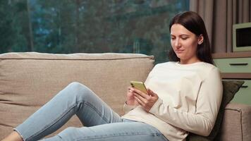 Woman relaxing on comfortable sofa, browsing social media using smartphone, enjoying leisure time in cozy home environment with soft natural light video