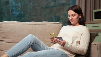 Happy customer woman is relaxing on comfortable sofa, using smartphone and credit card, buying online, making internet order, enjoying shopping at home video