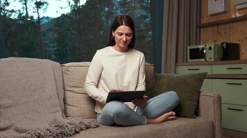 ung kvinna åtnjuter henne fritid tid bläddring på en digital läsplatta medan Sammanträde bekvämt på en soffa i en mysigt, modern Hem miljö med en se av de skog genom en stor fönster video