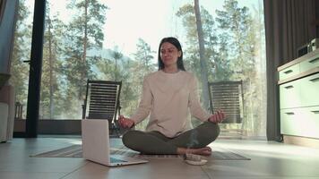 Relaxed woman enjoys a guided meditation session using her laptop, finding inner peace in her modern, stylish apartment with a large window overlooking a serene natural landscape video