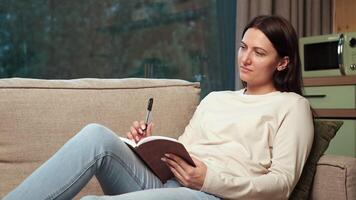 Young woman relaxing on comfortable living room sofa, thoughtfully writing in notebook while capturing personal reflections with creative concentration and peaceful introspection video