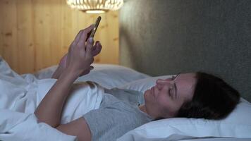Relaxed woman lying comfortably in bed during nighttime, engaging with smartphone while scrolling and tapping through digital content with soft illumination from screen video