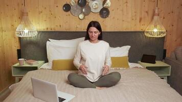 Serene woman practicing meditation while following online guidance, sitting in lotus position on bed within warm wooden bedroom interior, using laptop for wellness session video
