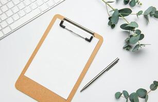 Clipboard with blank paper, pen, keyboard and eucalyptus branches on white table for working or studying photo
