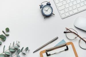Workspace organizing eucalyptus branches, keyboard, mouse, alarm clock, clipboard, pen, and eyeglasses on white table photo