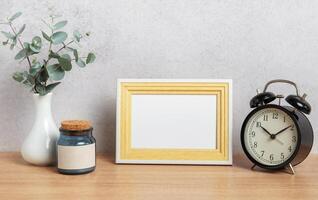 Empty photo frame, alarm clock, eucalyptus branches and jar on wooden table