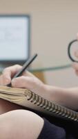 A person is focused on notetaking, adjusting their glasses with one hand while holding a pen, with a laptop in the background, showcasing a productive workspace full of creativity and learning video
