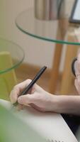 A closeup shows a hand gripping a sleek black pen, skillfully sketching in a spiral notebook. Elegant glass tables in the background contribute to a serene atmosphere for creativity video