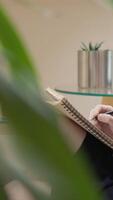A focused individual writes in an organized notebook on a sleek glass table, surrounded by lush plants and an elegant metal planter, creating a calm space ideal for creativity and mindfulness video