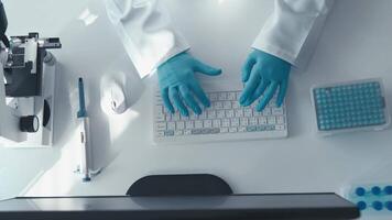 A top-down view of a laboratory workstation, featuring a researcher in blue gloves typing on a keyboard. Nearby are a microscope and lab equipment, reflecting a focused scientific environment. video