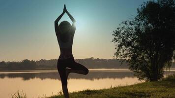 A womans silhouette gracefully practicing yoga at sunrise, harmonizing body and mind by a tranquil lakeside. The dawning light enhances the peaceful atmosphere, adding calm to the scene video