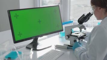 In a lab, a researcher in a white coat examines a sample under a microscope, while a nearby computer displays a bright green screen, showing an important, ongoing study in tech and science video