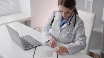 uma dedicada cuidados de saúde profissional dentro uma branco casaco é focado em escrevendo notas às uma escrivaninha enquanto a aberto computador portátil perto mostra seus comprometimento para diligente paciente Cuidado e manutenção de registros video