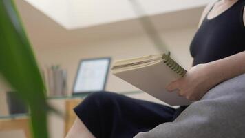 A person sits comfortably in a cozy chair, writing in a spiralbound notebook. They are in a serene indoor space with greenery and a computer, fostering inspiration and creativity video