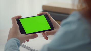 A person sits comfortably while holding a smartphone with a vibrant green screen. The cozy environment features soft lighting and blurred background elements, suggesting relaxation. video
