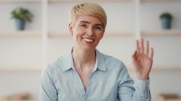 The woman is seated in a well-lit space, smiling and waving her hand to the camera, creating a friendly and welcoming atmosphere for viewers. video