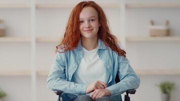 A cheerful girl with vibrant red hair is sitting in a wheelchair, smiling warmly as she engages in conversation. The background features a clean, modern indoor setting. video