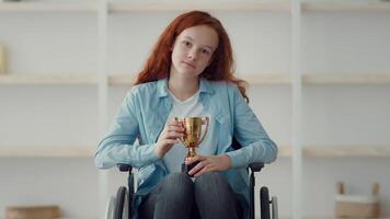 A girl with red hair sits in a wheelchair, proudly displaying a trophy. The bright room features shelves in the background, signifying her achievement and joy. video
