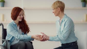 A caregiver gently holds the hands of a girl in a wheelchair. They share a moment of connection in a bright, soothing indoor space with soft lighting and minimal decor. video