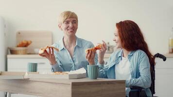 Mother and daughter share laughs and pizza while sitting at a wooden table in a bright, modern kitchen. The atmosphere is warm and inviting, highlighting their joyful friendship. video