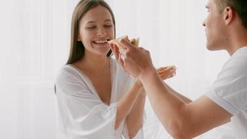 un joven Pareja disfruta un alegre momento en su acogedor dormitorio, alimentación cada otro Pizza con sonrisas, expresando su afecto y conexión en un relajado atmósfera. video