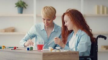 A woman and her daughter share a joyful moment, painting together at a wooden table filled with vibrant colors. They smile and engage, creating art in a well-lit, cozy space. video