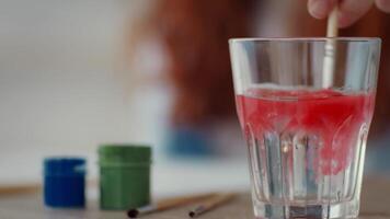 A person stirs vibrant red paint with a brush in a clear glass of water, surrounded by green and blue containers, creating a colorful setup for an artistic project. video