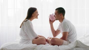 A young couple sits together on their bed in a bright bedroom, gazing into each other's eyes. The man holds a pink piggy bank, symbolizing their shared hopes for the future and financial goals. video