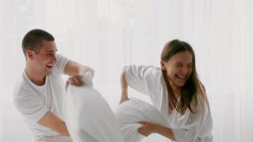 A young couple in a bright bedroom engages in a playful pillow fight. Their laughter fills the air as they share a moment of joy and affection together, creating a warm atmosphere. video