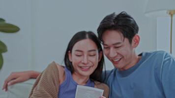 young couple sits on a sofa together, smiling and looking at the pregnancy results with joy, while their pug dog relaxes nearby. The bright room is filled with green plants creating a warm atmosphere. video