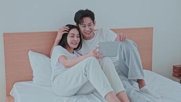 A young couple sits together on a bed, sharing laughter as they watch something engaging on a tablet. Their expressions reflect happiness and connection in a cozy home video