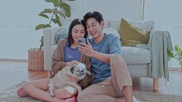 Couple enjoying sits comfortably on the floor of their modern living room, smiling as they interact with their smartphone while their dog rests between them, creating a cheerful atmosphere. video