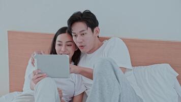 A young couple sits together on a bed, sharing laughter as they watch something engaging on a tablet. Their expressions reflect happiness and connection in a cozy home environment. video