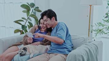 young couple sits on a sofa together smiling and looking at the pregnancy results with joy, while their pug dog relaxes nearby. The bright room is filled with green plants, creating a warm atmosphere. video