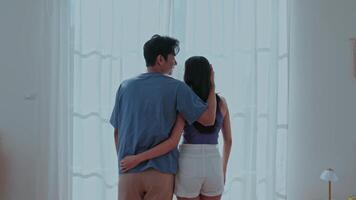 A couple stands close together in a sunny room, softly embracing. The light filters through sheer curtains, creating a warm atmosphere for their intimate moment. video