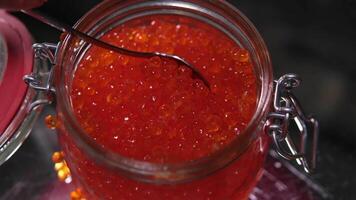Red Caviar in a spoon, fish roe in a glass jar. Close-up of salmon fish roe caviar on served table. Delicatessen. Texture of fresh trout caviar. Backdrop. Seafood. Slow motion. video