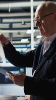 Vertical Smiling senior engineer in solar panels plant reading documentation to improve efficiency. Elderly man checking paperwork files in photovoltaics factory research and development department, camera A video