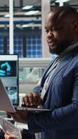 Vertical Portrait of smiling engineer in factory uses laptop to check parameters, improving solar panel durability. Happy African american researcher developing efficient sustainable energy solutions, camera A video