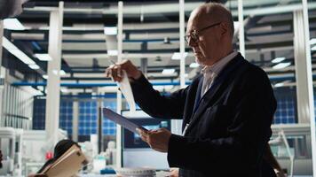 Smiling senior engineer in solar panels plant reading documentation to improve efficiency. Elderly man checking paperwork files in photovoltaics factory research and development department, camera A video