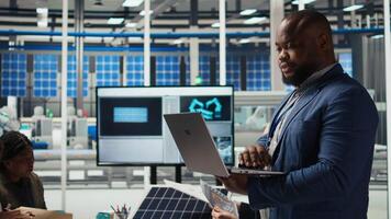 Portrait of smiling engineer in factory uses laptop to check parameters, improving solar panel durability. Happy African american researcher developing efficient sustainable energy solutions, camera A video