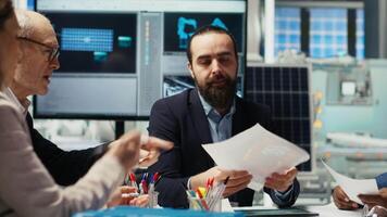 Business executives meet with investors to discuss the integration of automation technology in solar panel production processes, looking over paperwork and schematics, camera B video