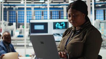 Portrait of researcher analyzes solar cell prototypes using laptop to optimize energy production. Smiling African american solar panels plant experts developing innovative manufacturing tech, camera A video